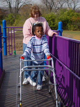 A child using a walker moves up the elevated ramp platform with assistance from an adult caregiver.