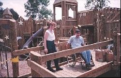Two people, a woman standing and a man using a wheelchair, are on an access ramp of a large wooden playground system