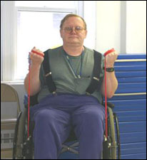 A man seated in a wheelchair is demonstrating the end position for a biceps curl exercise using a theraband