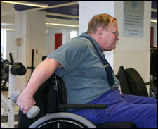 A man seated in a wheelchair is demonstrating the end position for a Double Arm Triceps Kickback with Free Weights exercise