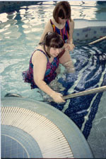 A young woman grips the handrail while using the steps to exit a pool. She receives assistance from a friend to sustain her balance and footing.