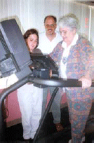 An older woman who has Alzheimer's Disease is exercising on a treadmill with the assistance of a staff and family member.