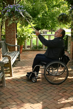 Wheelchair user tending hanging baskets with long garden tool. Pavers on ground are wheelchair accessible.