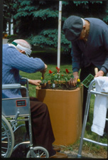 Two older adults, one standing and one using a wheelchair, tending a tall planter.