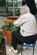 Older woman tending a tall planter while seated on a portable stool