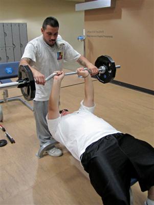 A man with a visual impairment has a spotter helping him while lifting weights.