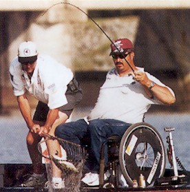 Two men are fishing: Man standing on the left holds net and man in wheelchair on the right holds fishing rod