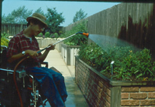 Young man using power wheelchair watering a brick raised bed against a fence 24 inched high and wide and 10 feet long.
