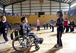 A girl, standing, plays catch with a boy in a wheelchair in a gym