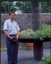 Man standing in front of a planting bed with green plants which encircles a round brick column.