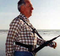 An older man walks near a lake with a harness rod holder attached to his body