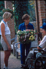 Two women talking to a seated older gentleman around a pot with a colorful bouquet