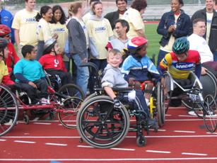 A boy in his manual wheelchair surrounded by wheelchair racers and others standing in the background on the track.