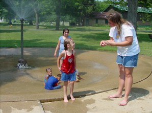Laurie is signing to a girl in front of a water sprinkler at a park.