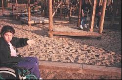 A young girl using a wheelchair sits in front of a wooden playground system surrounded by sand