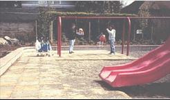 Two parents pushing their children on swings at a red playground with firm  and stable ground surfacing