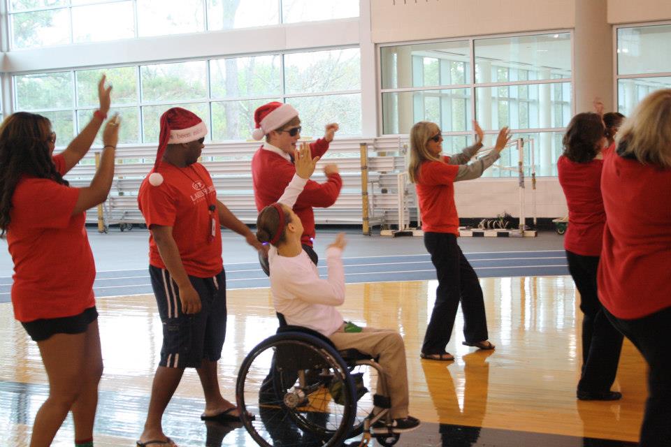 A group of people are wearing red shirts and santa hats and dancing in a gym.
