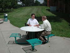 A young man in a wheelchair is seated at a round picnic table next to his friend.