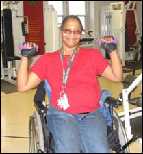 A woman seated in a wheelchair is demonstrating the start position for a Overhead Shoulder Press with Free Weights exercise.
