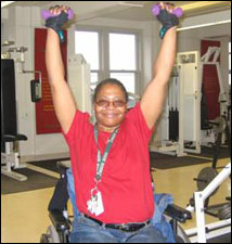 A woman seated in a wheelchair is demonstrating the end position for a Overhead Shoulder Press with Free Weights exercise.