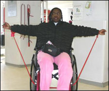 A woman seated in a wheelchair is demonstrating the end position for a Lateral Raise with Thera-Band® exercise.