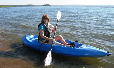 An adult woman with a physical disability is sitting in a t kayak on a lake.