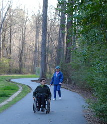 Image of two women on a path near a wooded area, one woman is walking while the other uses a wheelchair
