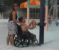 A mother stands behind son's wheelchair in shallow water at the water park.