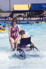 A woman stands near a woman using a wheelchair in a shallow pool.