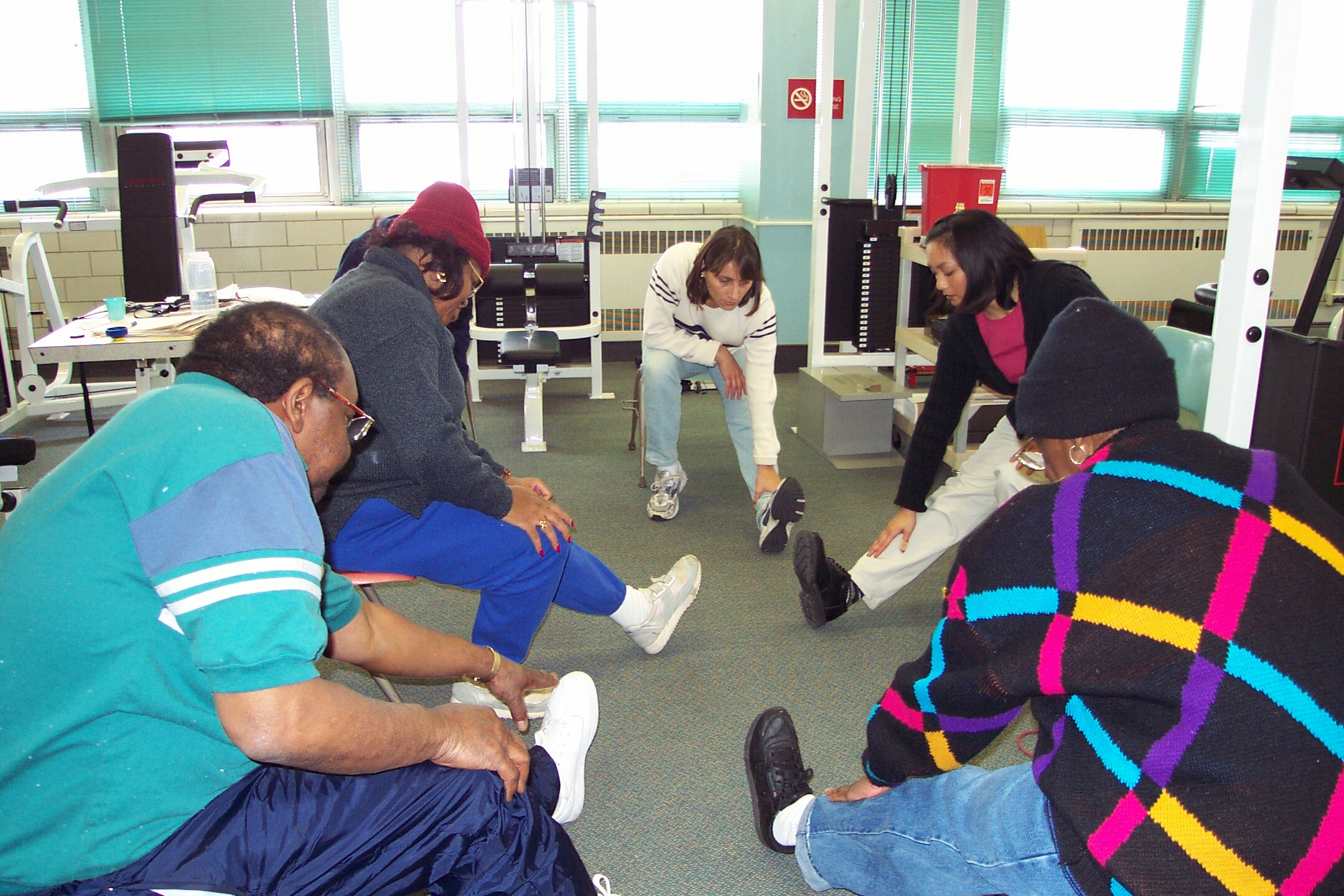 Five adults are seated are sitting in chairs in a circle leaning forward over an extended leg to stretch their hamstring.