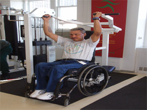 Man who is using a wheelchair is rolled into a shoulder press machine that has a swing-away seat