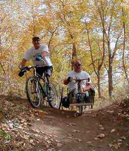Two men ride through the woods. One is riding a bike and the other is riding a handcycle.