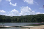 Blue sky with a few puffy white clouds over a lake surrounded by trees in the background and a dock in the foreground.