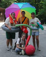 Male holding a brightly colored umbrella standing between two females with intertubes and another female sitting on the ground holding a red bat and a big red ball.