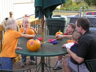 Two young girls, a boy, and a baby sitting on his father's lap decorate pumkins together on an outside table.