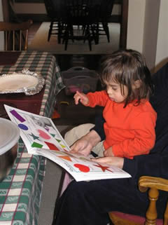 A young girl with downs syndrome points to a picture in a book, while sitting on her grandmothers lap.
