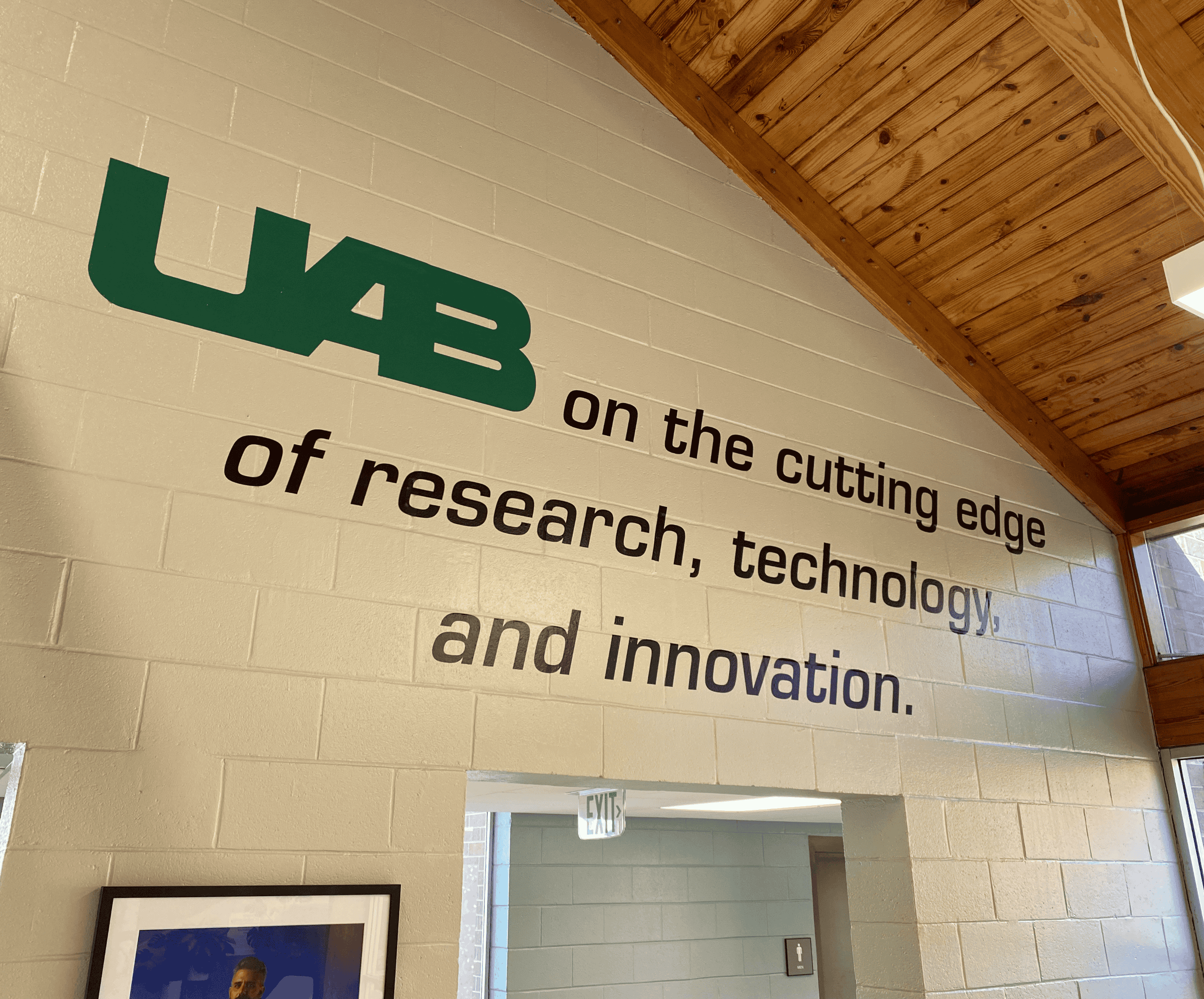 An interior wall with a slogan on it. It says "UAB on the cutting edge of research, technology, and innovation." It is above a doorway and below a wooden ceiling.